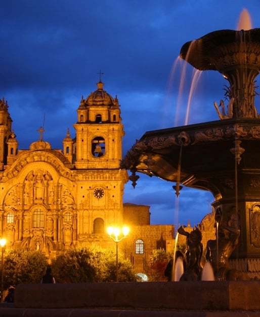 Colonial-style architecture of the Cusco Cathedral illuminated at night, with a decorative fountain in the foreground and a dramatic sky.