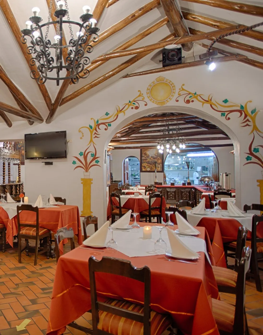 Interior view of Munay Wasi Inn's restaurant featuring colonial-style decor, wooden beams, vibrant wall art, and tables set with orange tablecloths, white napkins, and candles, highlighting the inviting dining atmosphere in Cusco, Peru.